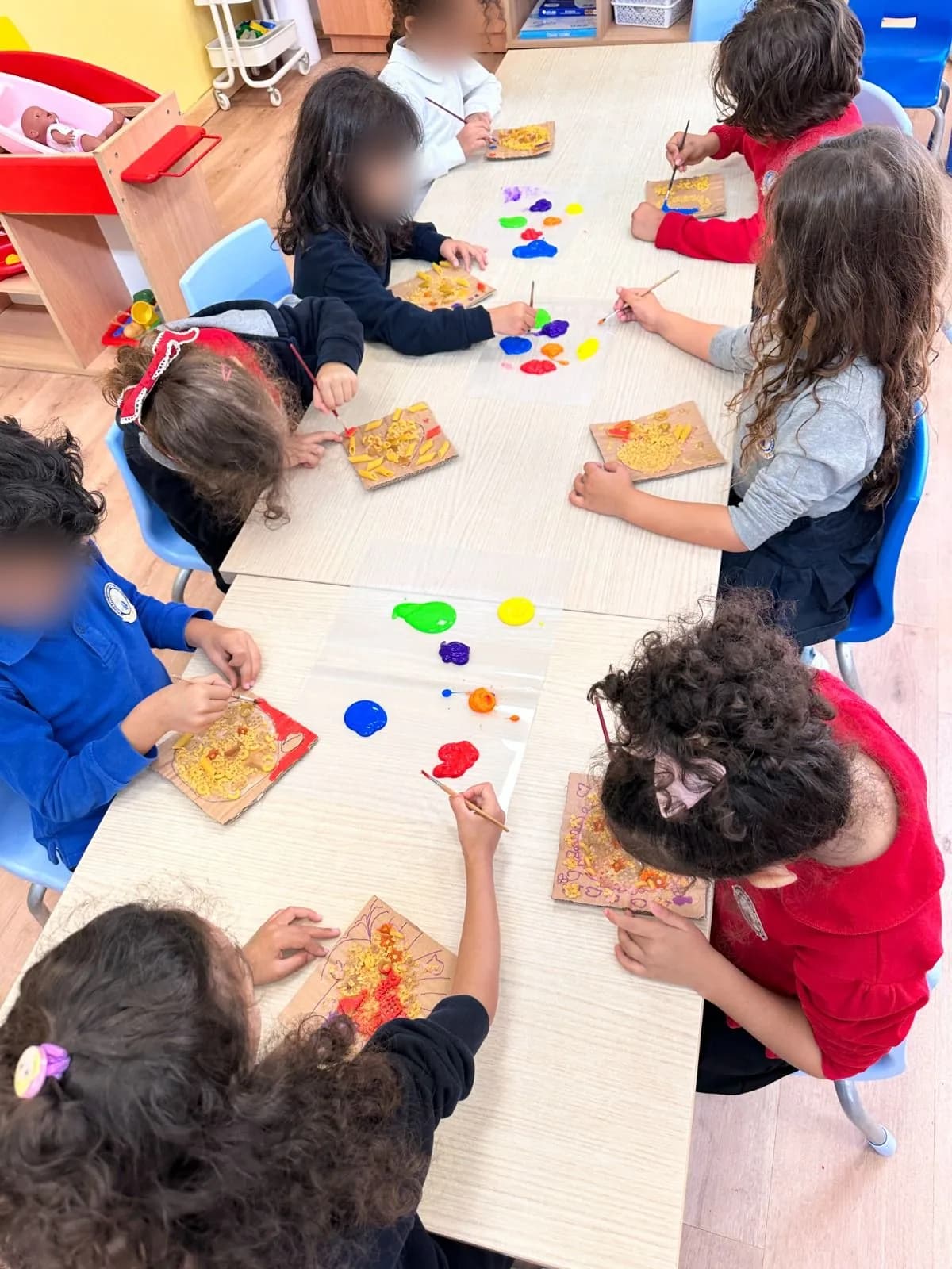Children painting pasta collages on cardboard around a long table
