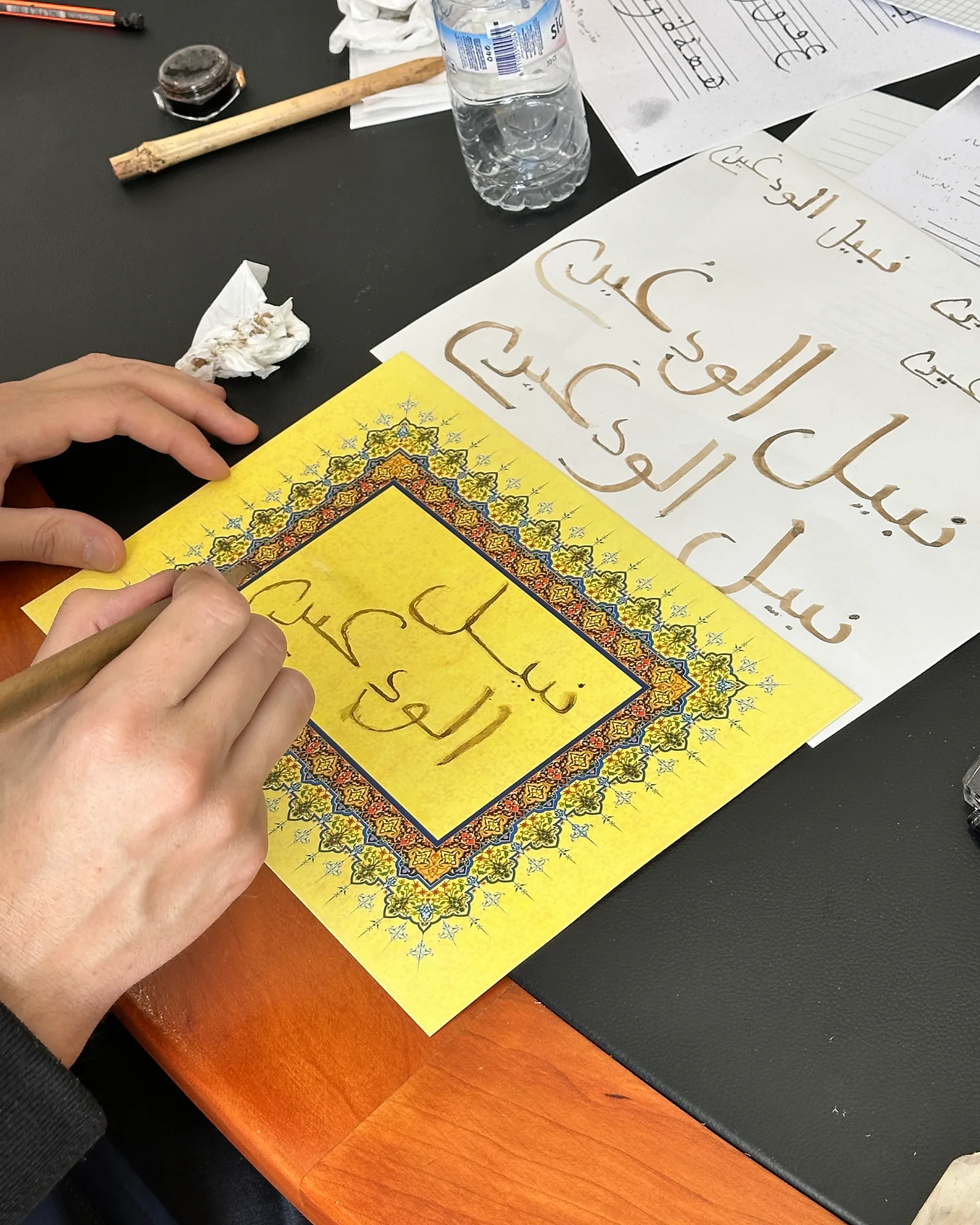 A hand writing Arabic calligraphy on an ornate card at Bluhimi Arts