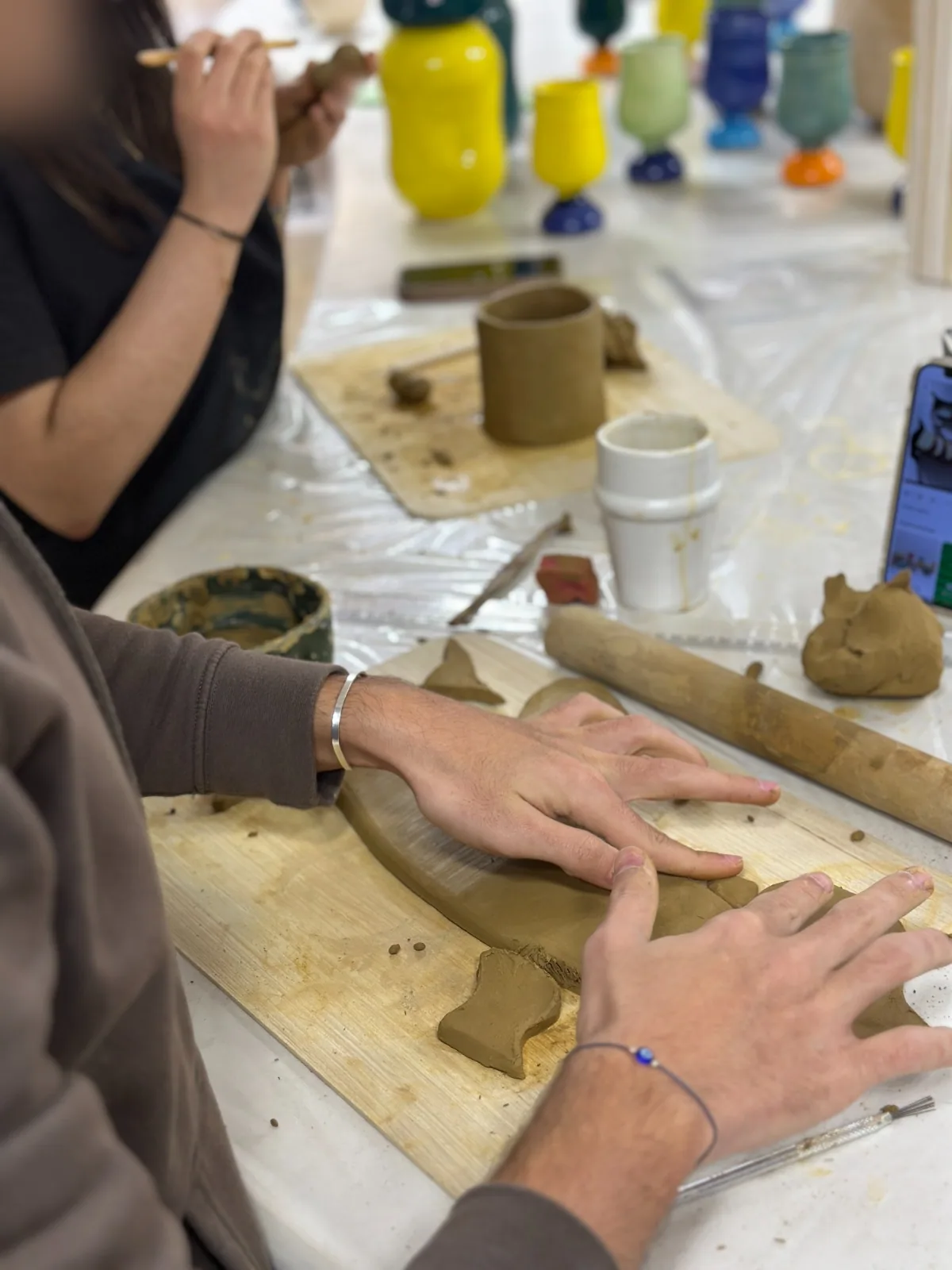 Hands pressing a slab of clay beside colourful ceramic vases
