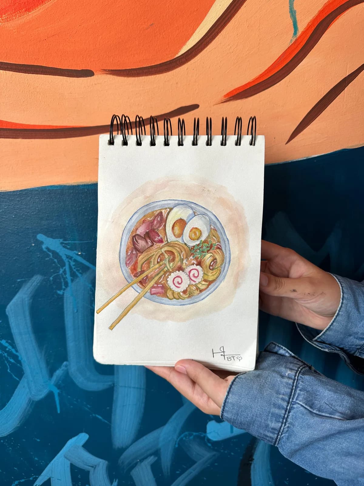 A watercolour sketch of a bowl of ramen held up against a painted wall